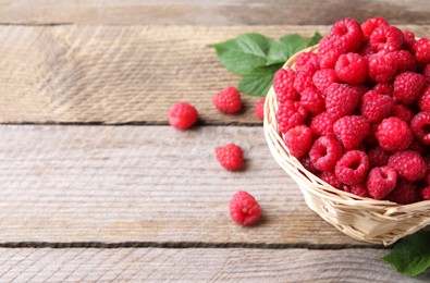Wicker basket with tasty ripe raspberries and green leaves on wooden table, space for text