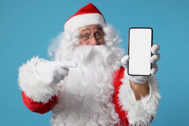 Photo of Merry Christmas. Santa Claus showing smartphone on light blue background, selective focus. Mockup for design