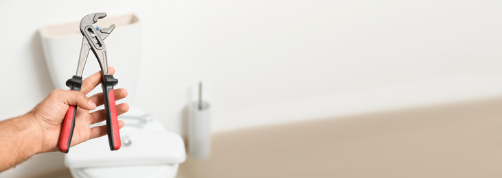 Closeup view of professional plumber holding pliers against toilet bowl in bathroom, space for text. Banner design