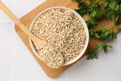 Photo of Dry pearl barley in bowl, spoon and parsley on white tiled table, top view