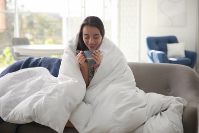 Photo of Beautiful young woman wrapped with soft blanket holding cup of hot drink on sofa at home