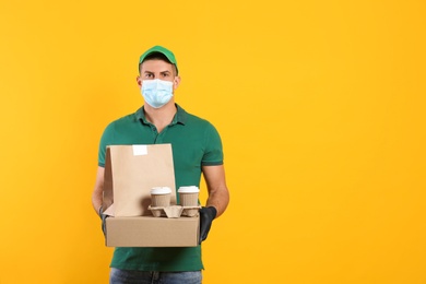 Photo of Courier in medical mask holding packages with takeaway food and drinks on yellow background, space for text. Delivery service during quarantine due to Covid-19 outbreak