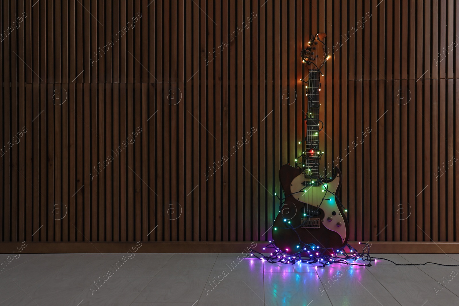 Photo of Modern electric guitar with Christmas lights on floor near wooden wall. Space for text