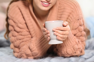 Attractive smiling young woman in cozy warm sweater with cup of hot drink lying on floor at home