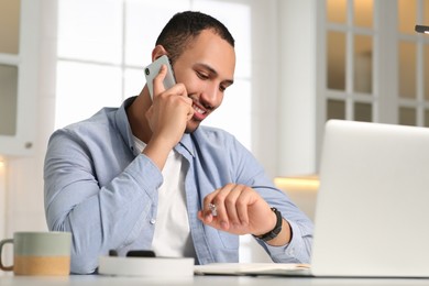 Young man talking on smartphone while working with laptop at desk in kitchen. Home office