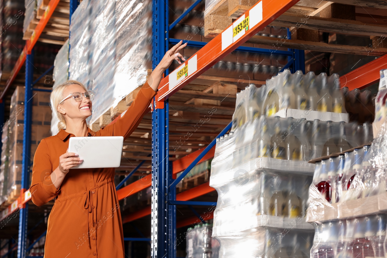 Photo of Happy manager holding modern tablet and pointing at something in warehouse with lots of products