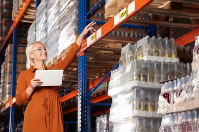 Photo of Happy manager holding modern tablet and pointing at something in warehouse with lotsproducts