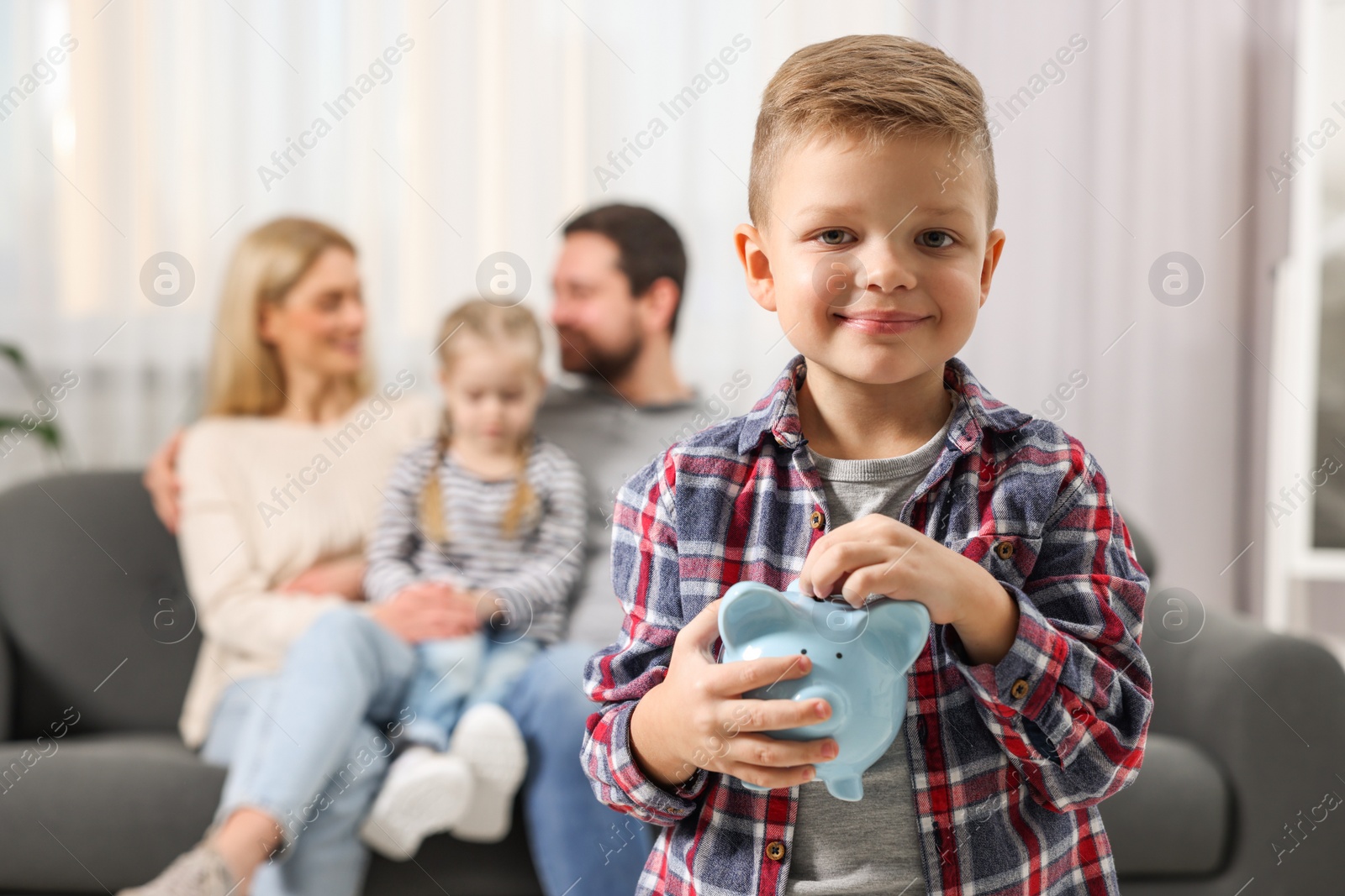 Photo of Family budget. Little boy putting coin into piggy bank while his parents and sister sitting on sofa at home, selective focus