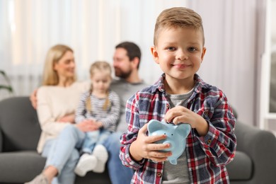 Photo of Family budget. Little boy putting coin into piggy bank while his parents and sister sitting on sofa at home, selective focus