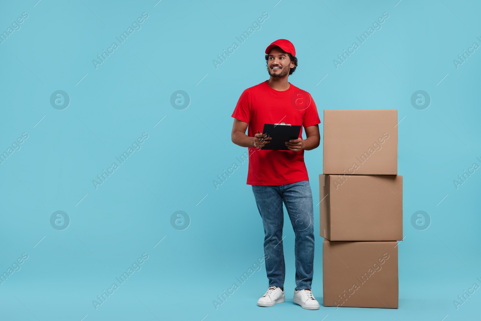 Photo of Happy young courier with clipboard and stack of parcels on light blue background, space for text