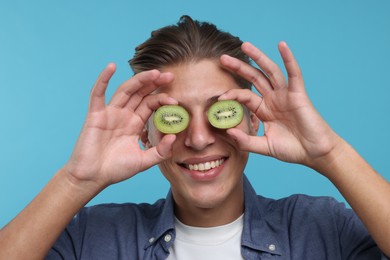 Photo of Smiling man covering eyes with half of kiwi on light blue background, closeup
