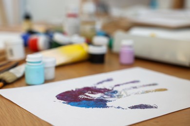 Photo of Sheet of paper with handprint made with colorful paints on wooden table indoors, closeup. Creative hobby