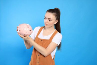 Sad young woman with piggy bank on light blue background