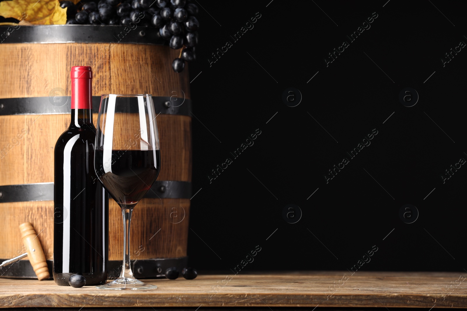 Photo of Delicious wine, wooden barrel and ripe grapes on table against black background. Space for text