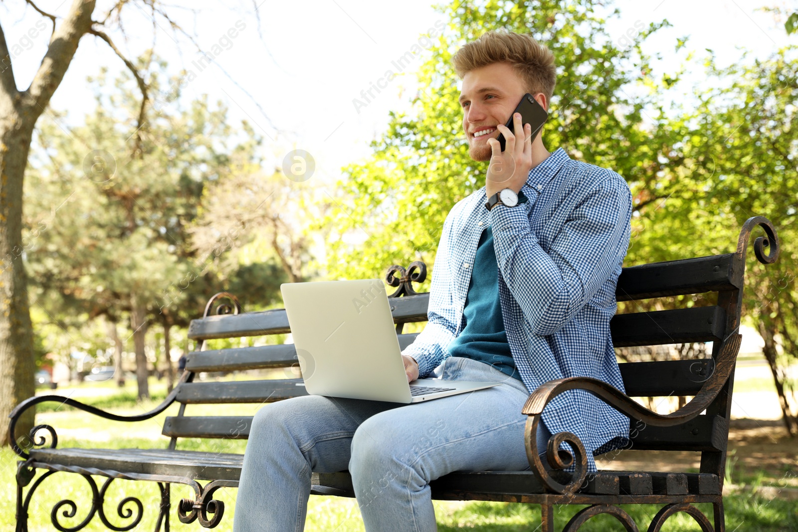 Image of Happy young man with laptop talking on phone in park