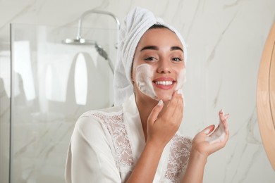 Photo of Beautiful young woman applying cleansing foam onto face in bathroom. Skin care cosmetic
