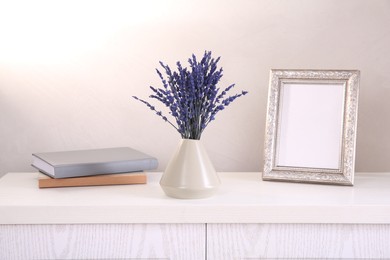 Bouquet of beautiful preserved lavender flowers, notebooks and blank photo frame on white wooden chest of drawers near beige wall