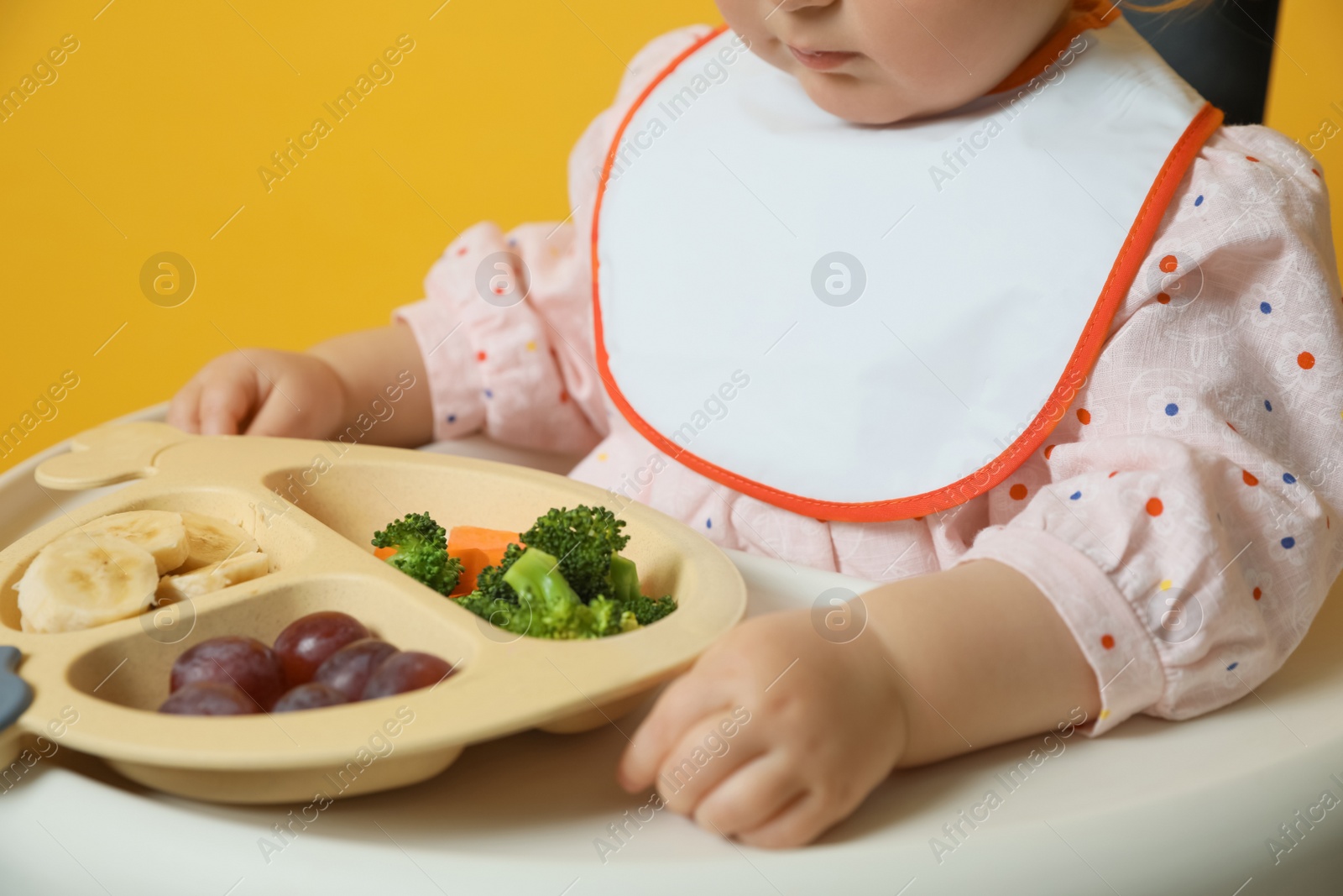 Photo of Cute little baby wearing bib while eating on yellow background, closeup