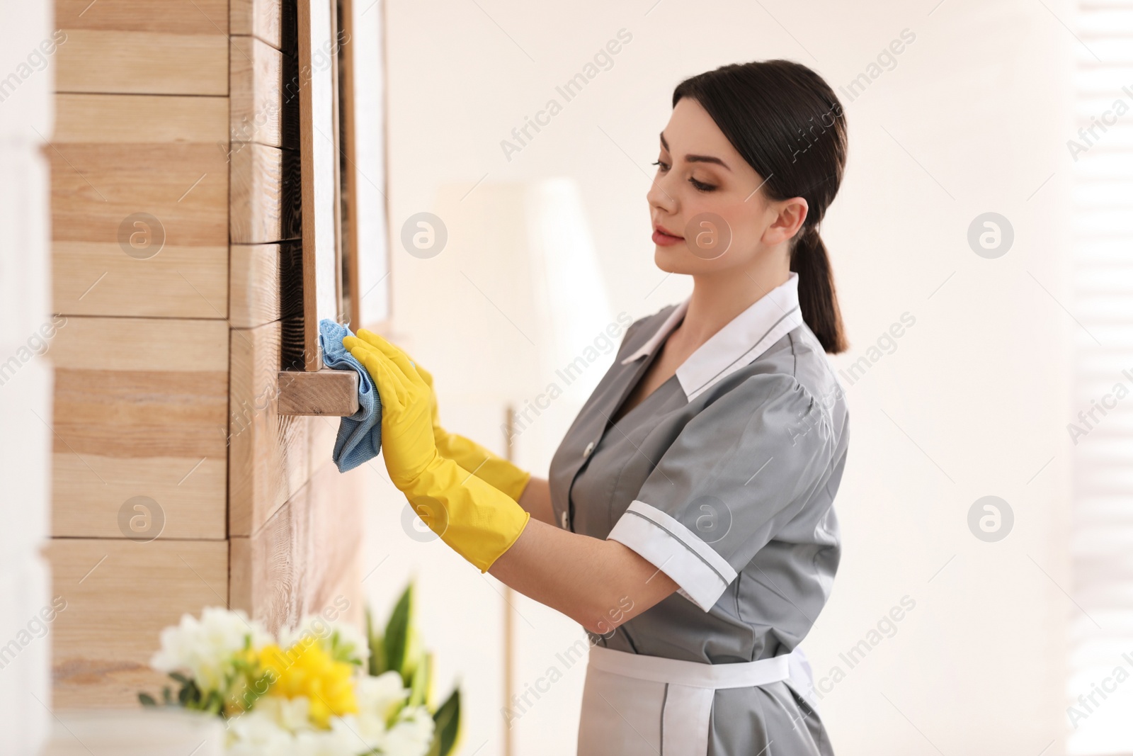 Photo of Young chambermaid wiping dust from furniture in hotel room