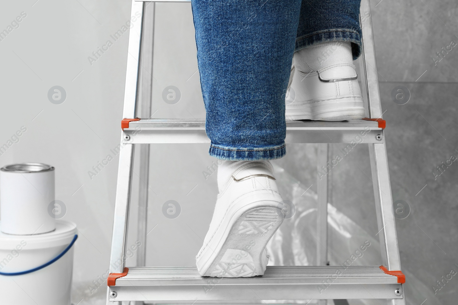 Photo of Woman climbing metallic folding ladder, can and bucket of paint near grey wall, closeup