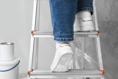 Photo of Woman climbing metallic folding ladder, can and bucket of paint near grey wall, closeup