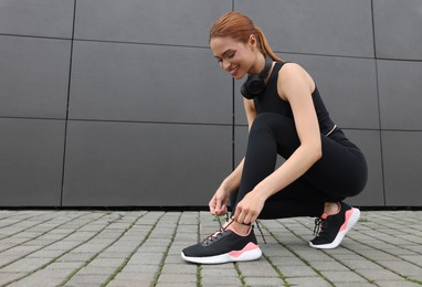 Beautiful woman in stylish gym clothes with headphones tying shoelace of sneakers near dark grey wall on street, space for text
