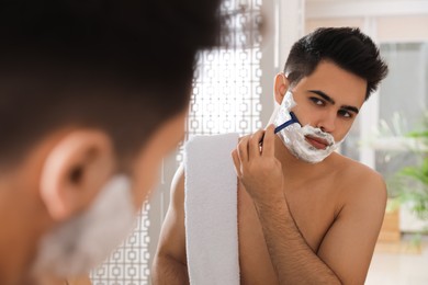 Photo of Handsome young man shaving with razor near mirror in bathroom