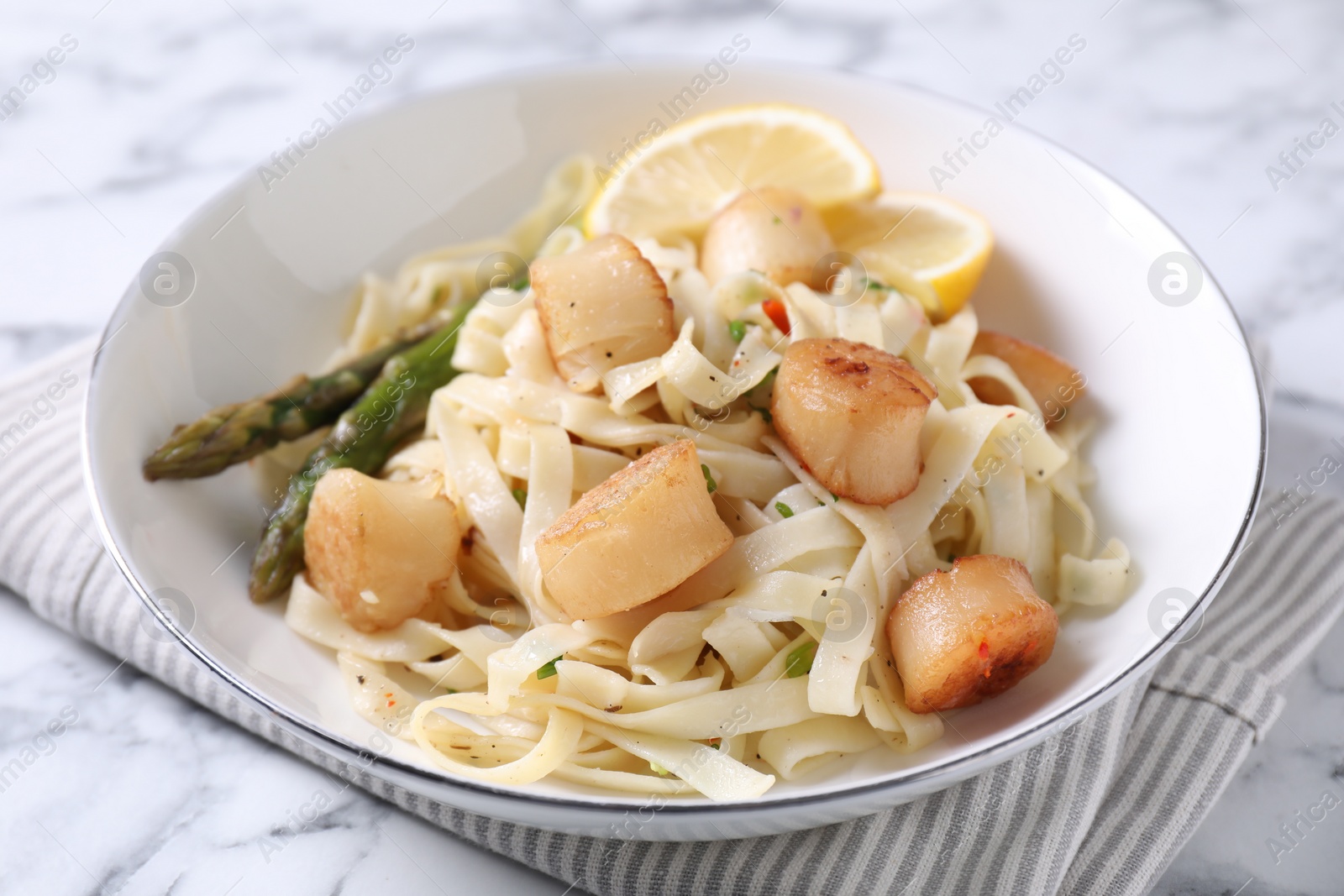 Photo of Delicious scallop pasta with asparagus and lemon on white marble table, closeup