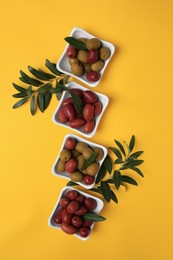 Different fresh olives and green leaves on yellow background, flat lay