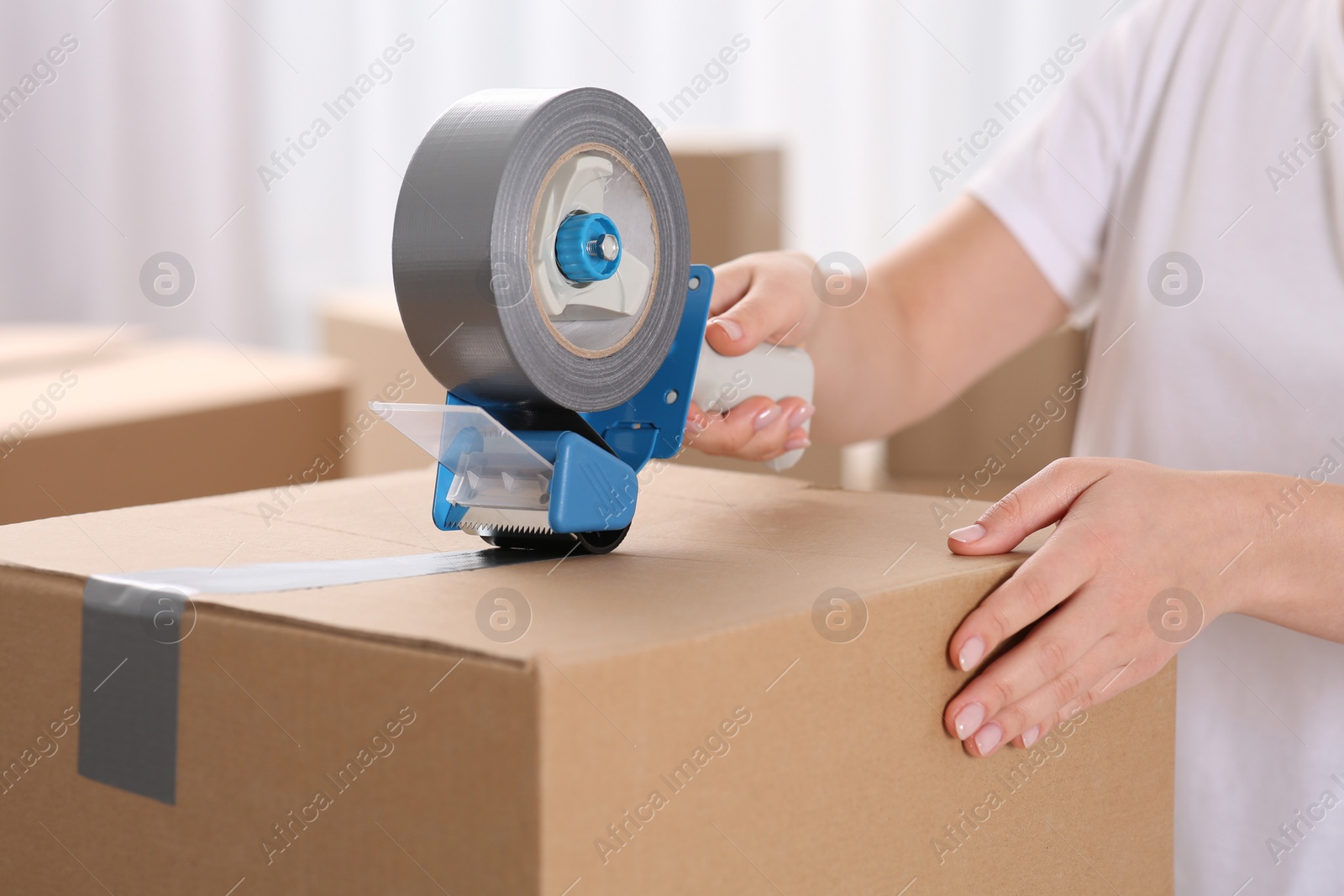 Photo of Woman applying adhesive tape on box with dispenser indoors, closeup