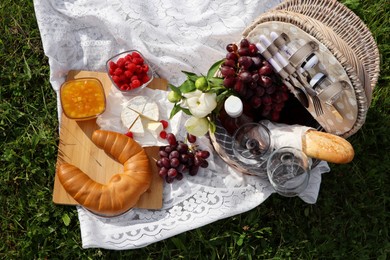 Picnic blanket with tasty food, flowers, basket and cider on green grass outdoors, flat lay