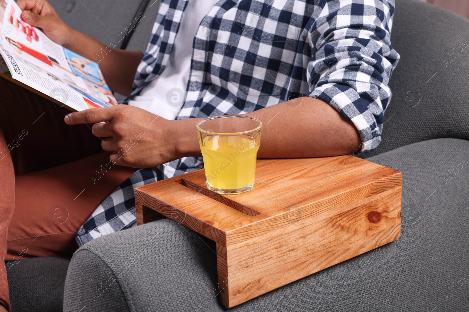 Photo of Glass of juice on wooden sofa armrest table. Man reading magazine at home, closeup