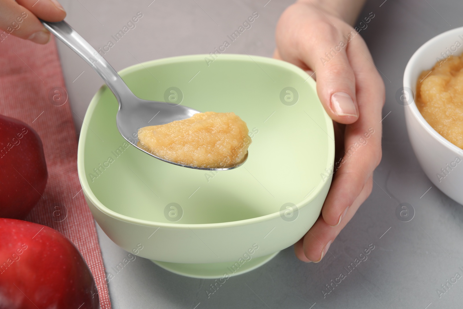 Photo of Healthy baby food. Woman putting delicious apple puree into bowl at light grey table, closeup