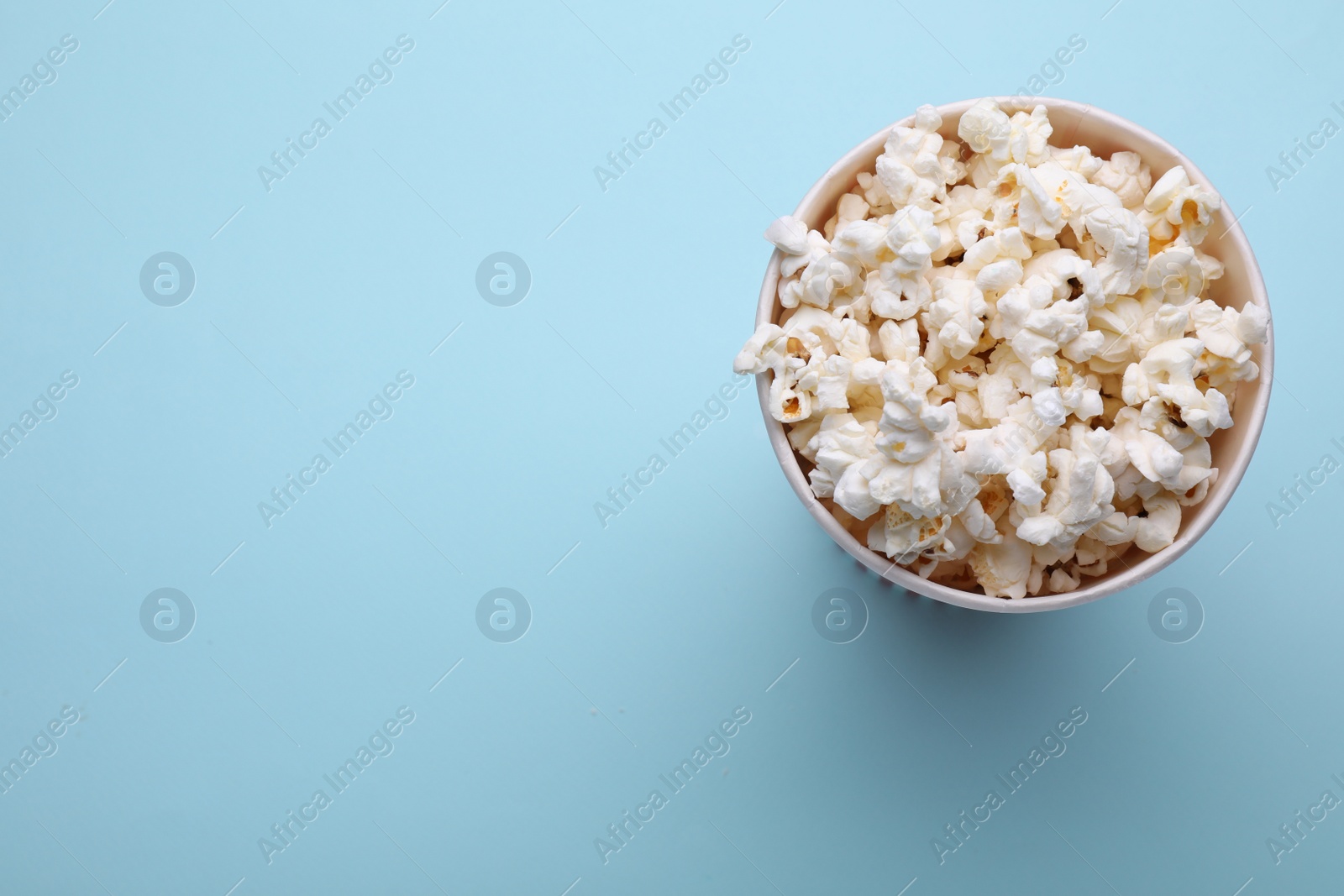 Photo of Paper bucket with delicious popcorn on light blue background, top view. Space for text