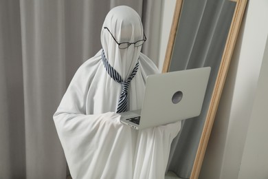 Photo of Creepy ghost. Man in white sheet using laptop at home