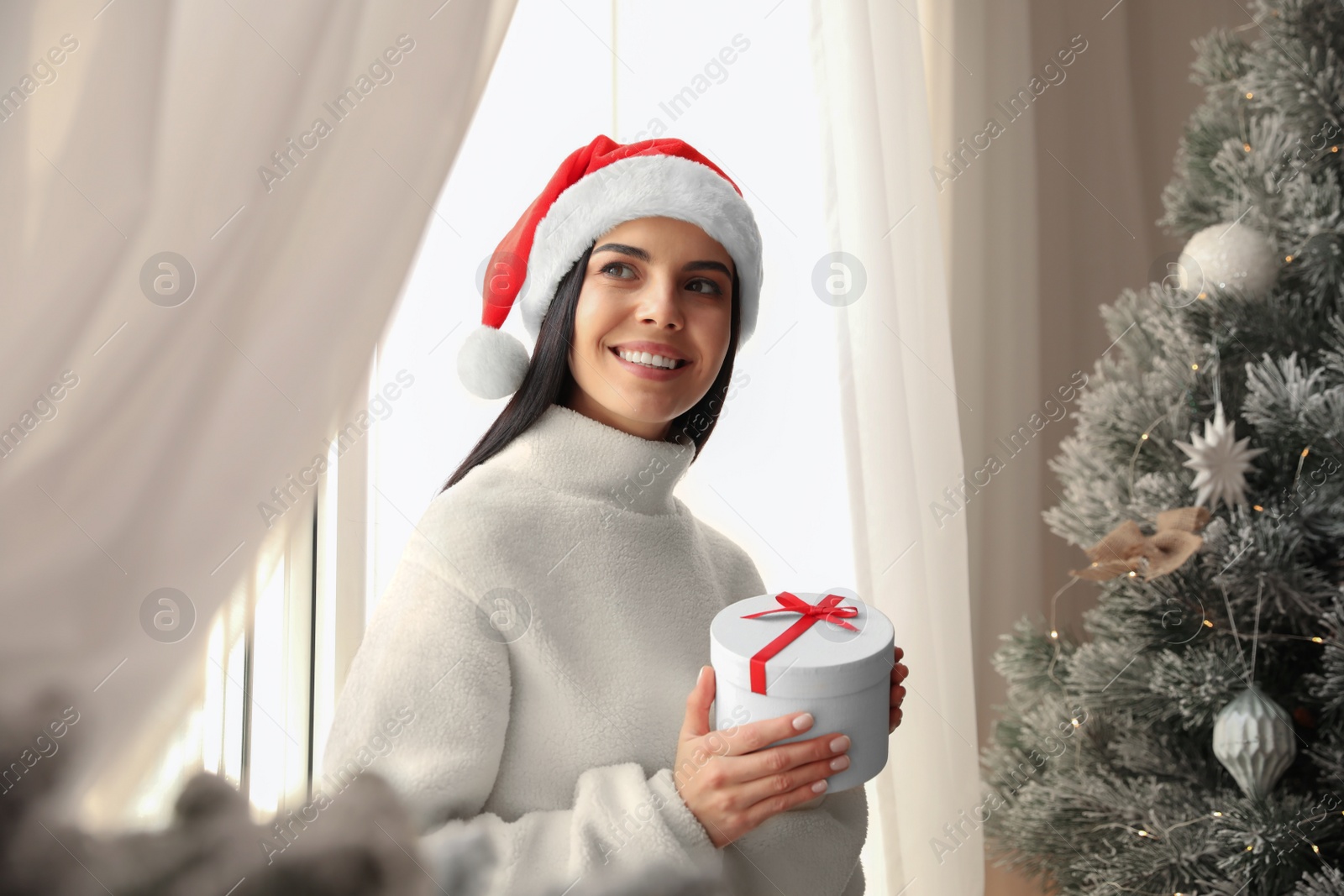 Photo of Woman in Santa hat holding gift box near Christmas tree