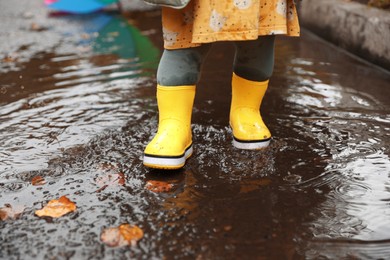 Photo of Little girl splashing water with her boots in puddle outdoors, closeup