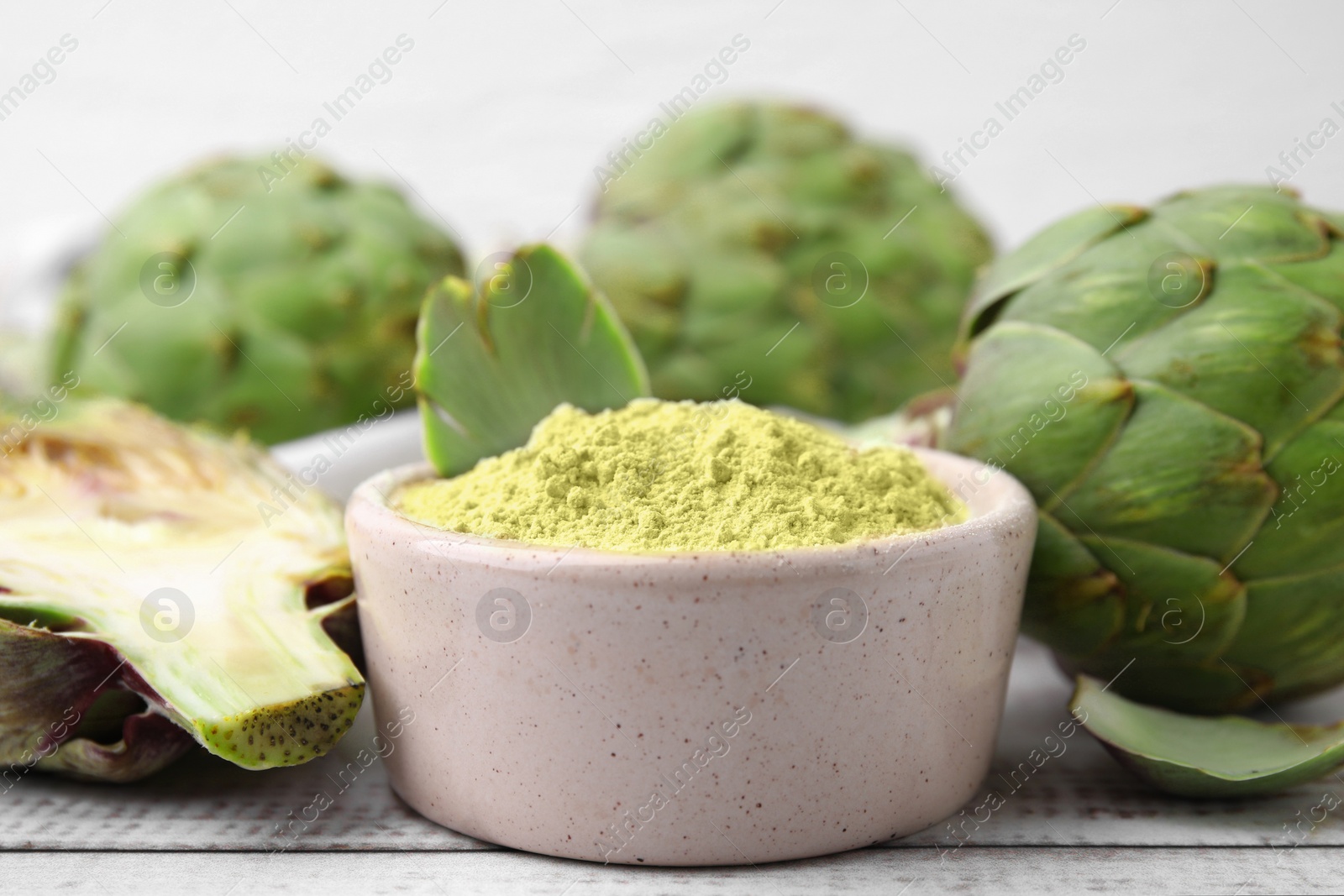 Photo of Bowl with powder and fresh artichokes on white wooden table, closeup