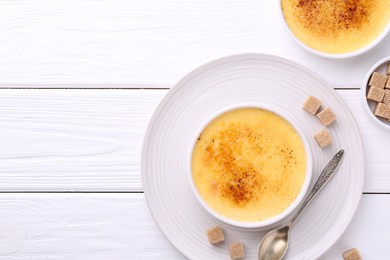 Photo of Delicious creme brulee in bowls, sugar cubes and spoon on white wooden table, flat lay. Space for text