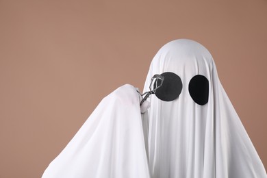 Photo of Fashionable ghost. Woman covered with white sheet using eyelash curler on dark beige background, space for text