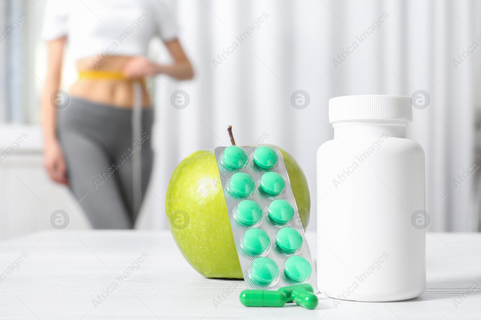 Photo of Weight loss pills with apple on table and woman measuring waist indoors