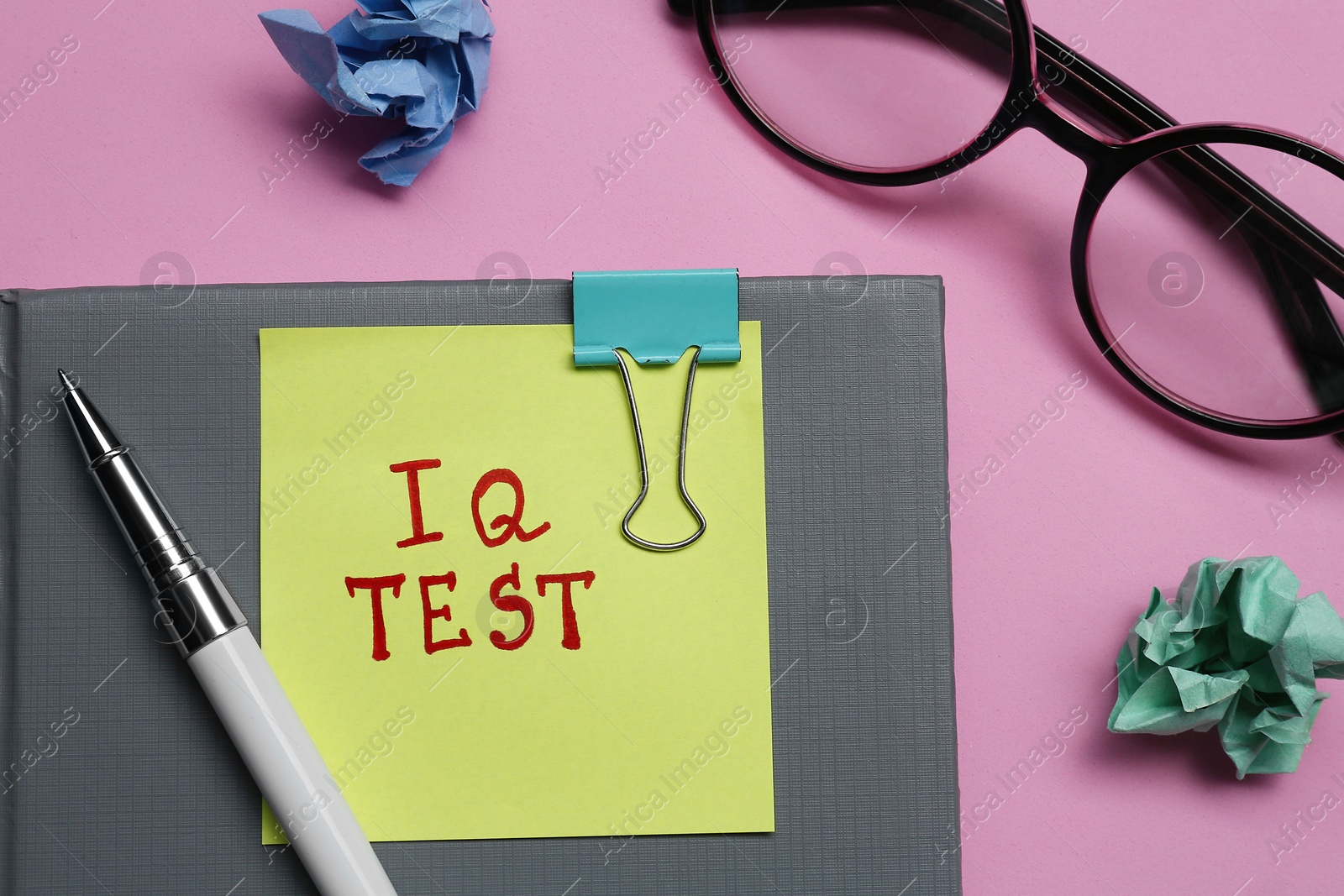 Photo of Sticker note with text IQ Test, notebook, pen, crumpled paper balls and glasses on pink background, flat lay