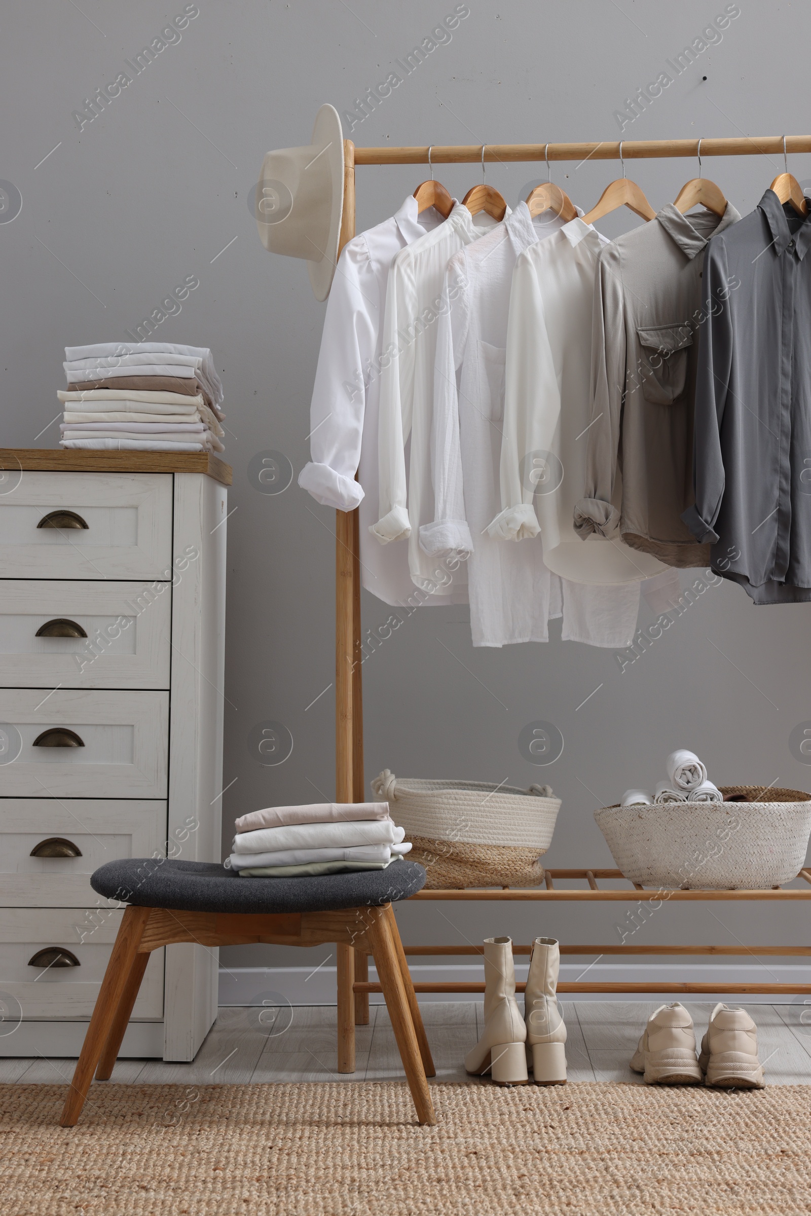 Photo of Wardrobe organization. Rack with different stylish clothes, accessories, ottoman and chest of drawers near grey wall indoors