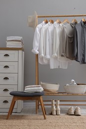 Wardrobe organization. Rack with different stylish clothes, accessories, ottoman and chest of drawers near grey wall indoors
