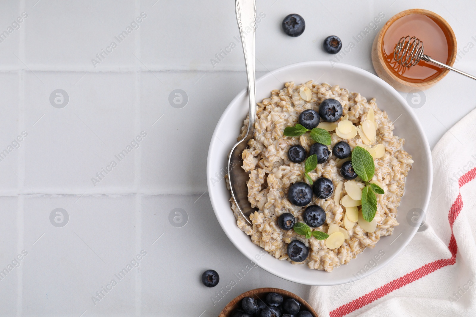 Photo of Tasty oatmeal with blueberries, mint and almond flakes in bowl on white tiled table, flat lay. Space for text