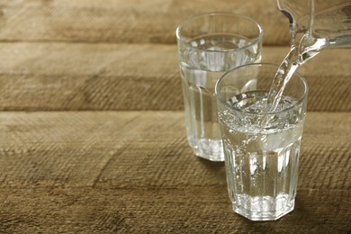 Photo of Pouring water from jug into glass on wooden table, space for text