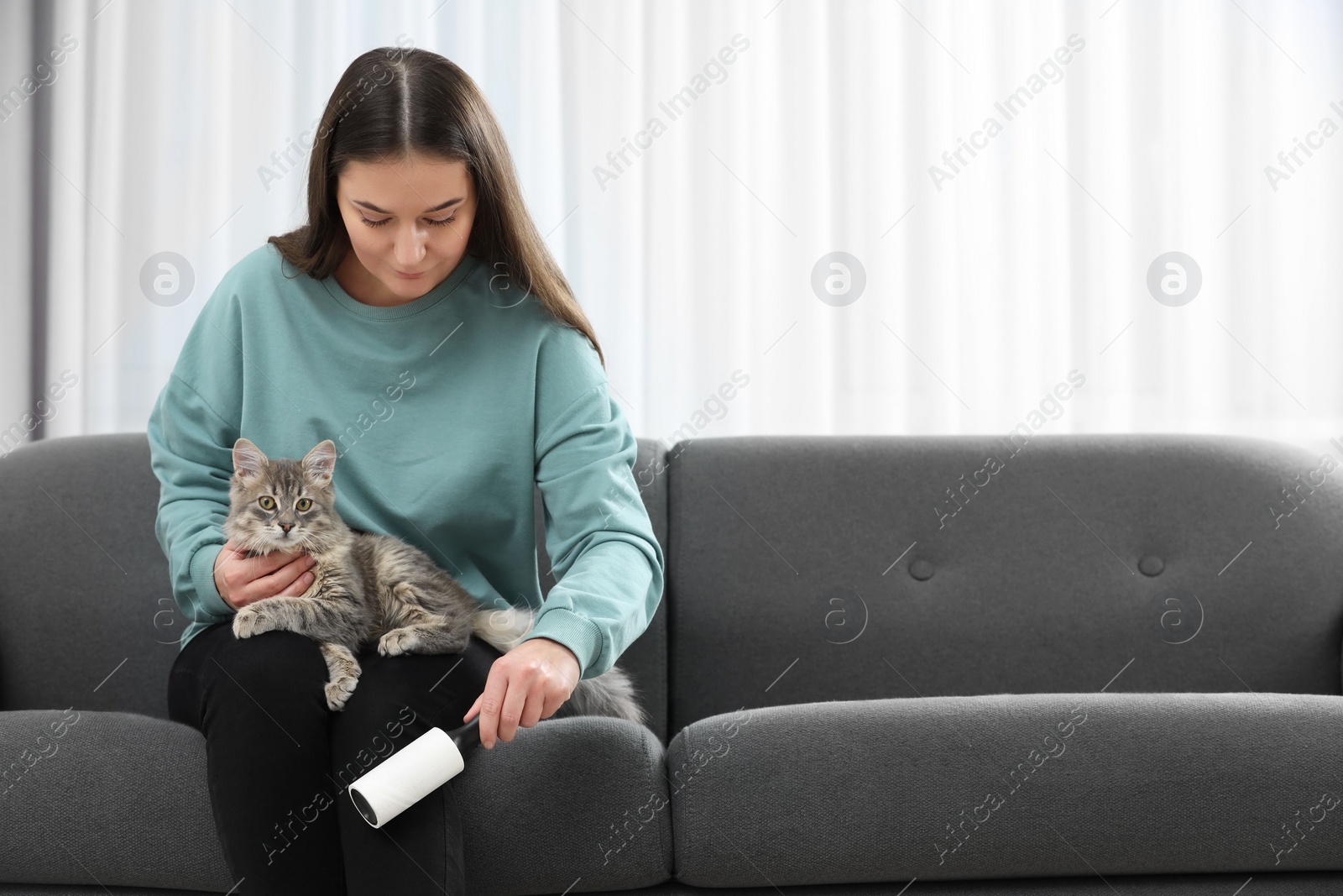 Photo of Pet shedding. Woman with lint roller removing cat`s hair from trousers on sofa at home, space for text