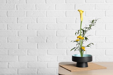 Photo of Beautiful ikebana for stylish house decor. Floral composition with fresh calla, chrysanthemum flowers and branches on wooden table near white brick wall, space for text