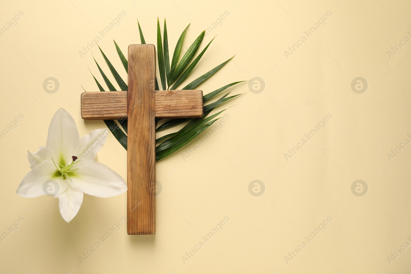 Photo of Wooden cross, lily flower and palm leaf on pale yellow background, top view with space for text. Easter attributes