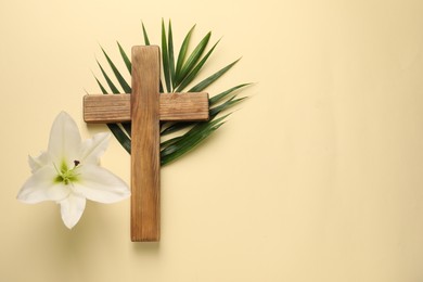 Wooden cross, lily flower and palm leaf on pale yellow background, top view with space for text. Easter attributes